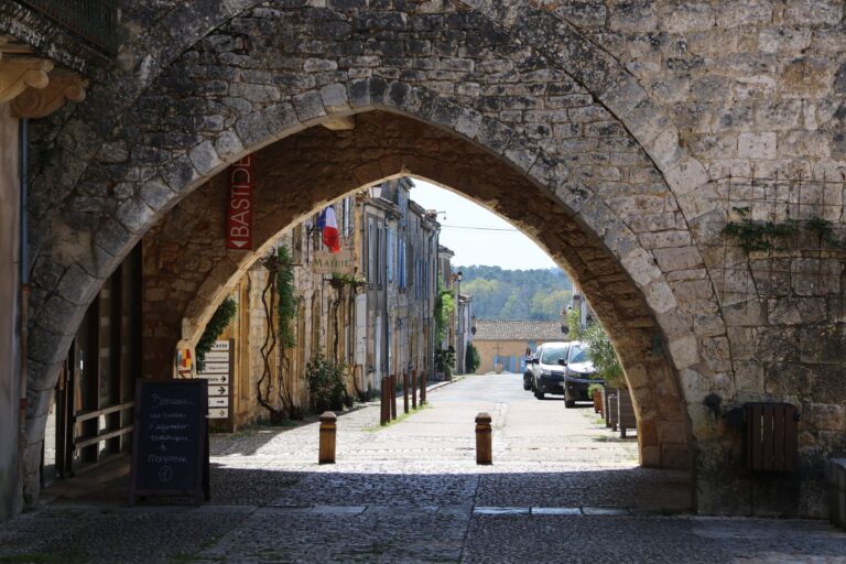 Porte fortifiée en pierre dorée et ruelle pavée d'une bastide du Sud-Ouest en fin d'été